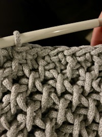 a person knitting a grey crochet stitch with a white crochet hook